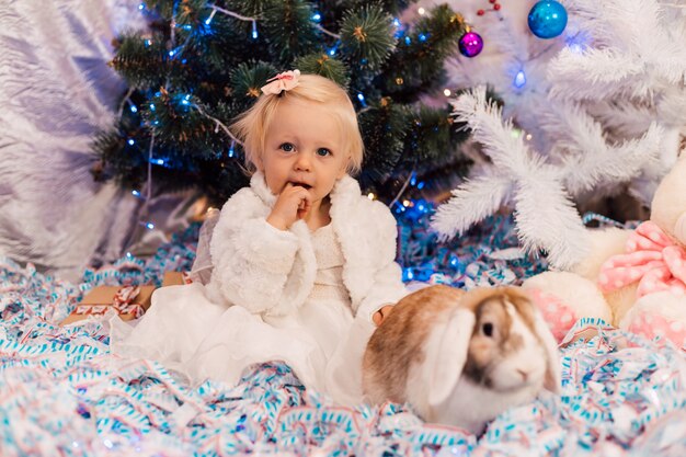 menina perto de uma árvore de natal com um vestido beome brincando com um coelho