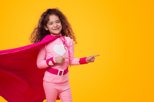 Foto menina positiva com fantasia de super-herói rosa com capa voadora, olhando para a câmera com um sorriso e apontando contra um fundo amarelo