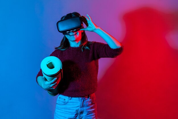 Menina positiva com óculos de realidade virtual segurando um tapete de ioga enrolado