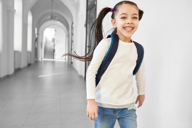 Menina positiva e feliz da escola que corre para dirigir do corredor e do sorriso da escola.