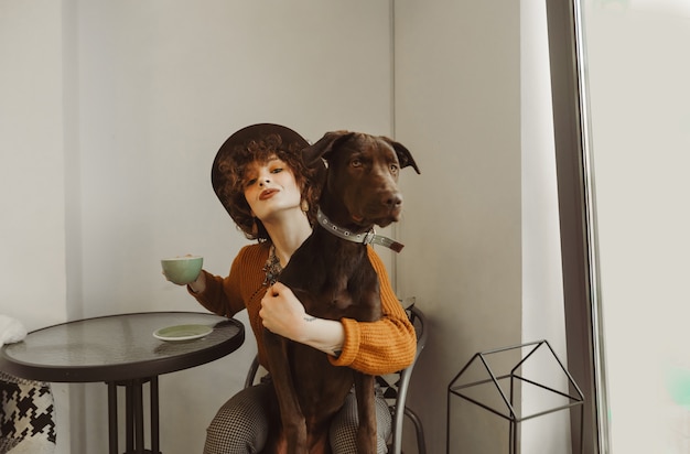 Menina positiva em roupas elegantes e chapéu sentado à mesa com uma xícara de café na mão