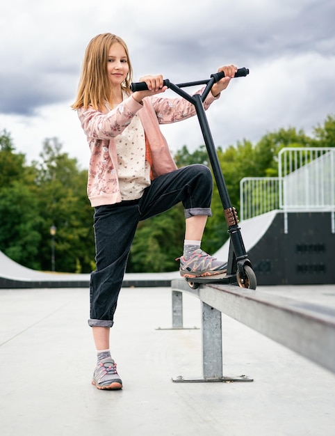 Menina pré-adolescente em pé com scooter na rua no parque e olhando para a câmera linda criança posando sagacidade