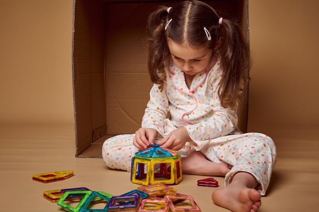 Menina pré-escolar concentrada de pijama sentada à frente de uma caixa de papelão e brincando com o construtor magnético entretenimento infantil e desenvolvimento motor fino mata