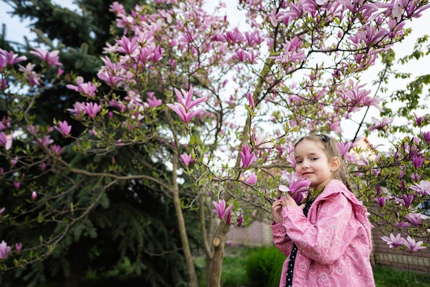 Menina pré-escolar em jaqueta rosa aproveitando o belo dia de primavera perto da árvore de magnólia florescendo Atividades de primavera