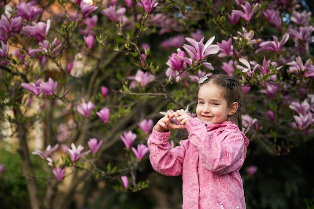 Menina pré-escolar em jaqueta rosa mostrando o coração pelos dedos enquanto desfruta de um belo dia de primavera perto da árvore florescente de magnólia