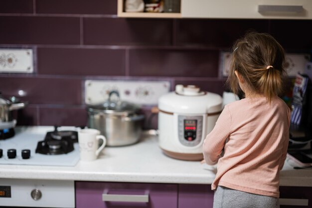 Menina prepara o jantar na cozinha de casa com fogão lento e libera vapor do vaporizador