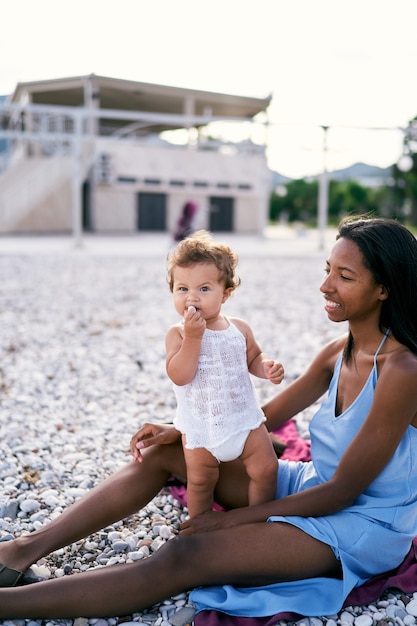 Menina roendo seixos ao lado da mãe sentada na praia
