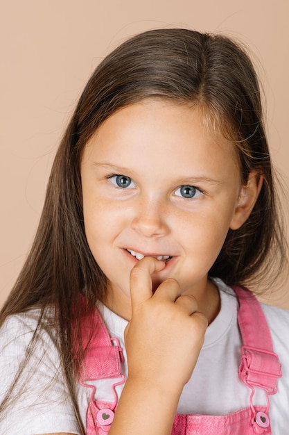 Menina roendo unhas com olhos brilhantes sorrindo olhando para a câmera vestindo macacão rosa brilhante e camiseta branca em fundo bege