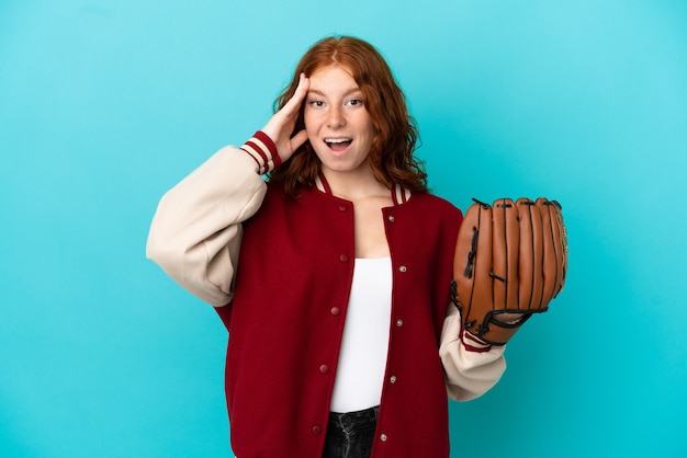 Menina ruiva adolescente com luva de beisebol isolada em um fundo azul com expressão de surpresa
