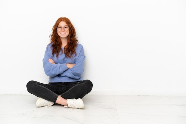 Menina ruiva adolescente sentada no chão, isolada no fundo branco, com os braços cruzados e olhando para a frente