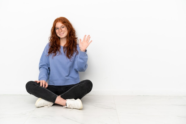 Menina ruiva adolescente sentada no chão, isolada no fundo branco, saudando com a mão e com uma expressão feliz