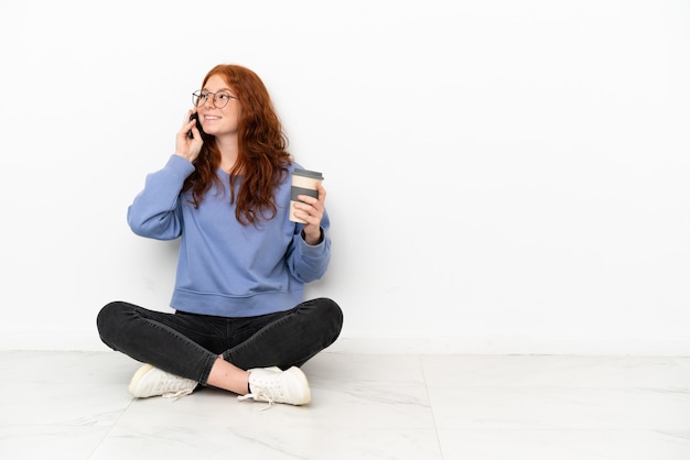 Menina ruiva adolescente sentada no chão, isolada no fundo branco, segurando um café para levar e um celular