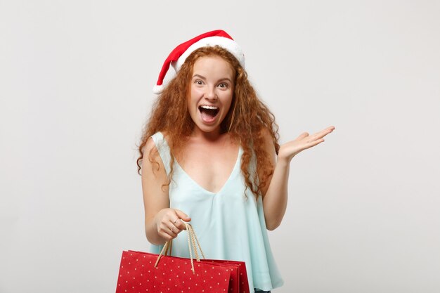 Menina ruiva de papai noel animado com chapéu de natal, isolado no fundo branco. feliz ano novo conceito de feriado de celebração de 2020. simule o espaço da cópia. segure a sacola com presentes ou compras após as compras.