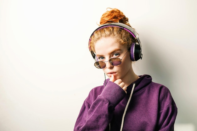 Menina ruiva encaracolada em fones de ouvido usando óculos e capuz roxo ouvindo sua música favorita