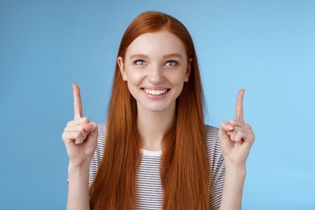 Menina ruiva sorridente, divertida e animada, olhando para cima sorrindo de bom grado assistindo a incrível performance ao ar livre em pé, mostrando uma coisa legal indicando o produto, fundo azul