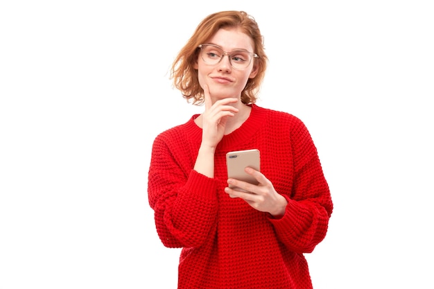 Menina ruiva sorridente pensativa segurando o smartphone nas mãos com óculos e roupas vermelhas isoladas no fundo branco do estúdio. Recebeu mensagem alegre
