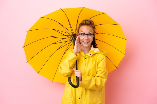 Menina russa adolescente com casaco à prova de chuva e guarda-chuva isolado em fundo rosa com surpresa e expressão facial chocada