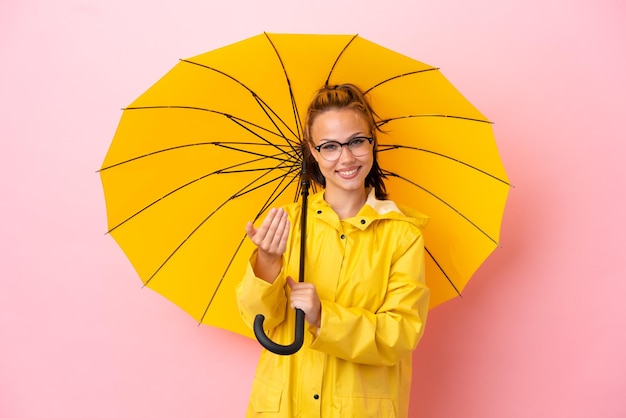 Menina russa adolescente com casaco à prova de chuva e guarda-chuva isolado em fundo rosa convidando para vir com a mão Feliz que você veio