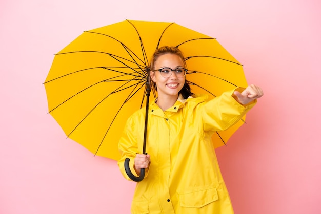Menina russa adolescente com casaco à prova de chuva e guarda-chuva isolado em fundo rosa dando um polegar para cima gesto