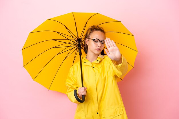 Menina russa adolescente com casaco à prova de chuva e guarda-chuva isolado em fundo rosa fazendo gesto de parada e desapontado