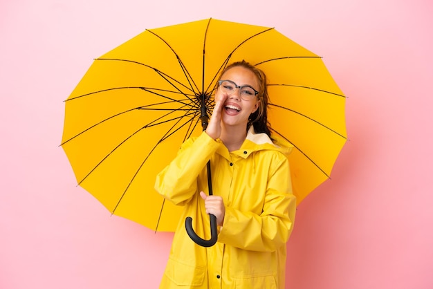 Menina russa adolescente com casaco à prova de chuva e guarda-chuva isolado em fundo rosa gritando com a boca aberta