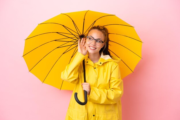 Menina russa adolescente com casaco à prova de chuva e guarda-chuva isolado em fundo rosa ouvindo algo colocando a mão na orelha