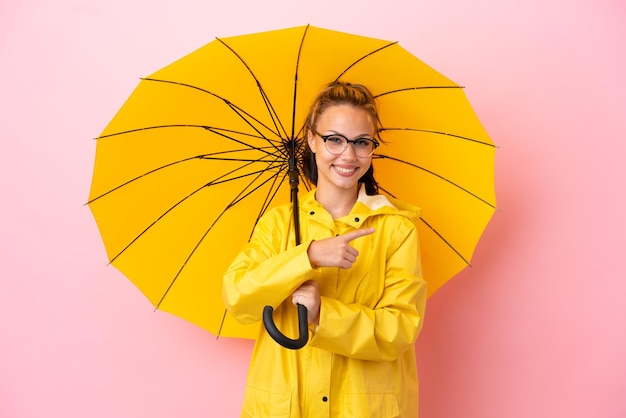 Menina russa adolescente com casaco à prova de chuva e guarda-chuva isolado em um fundo rosa apontando para trás
