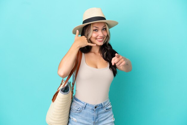 Menina russa adolescente com pamel e bolsa de praia isolada em fundo azul, fazendo gesto de telefone e apontando para a frente