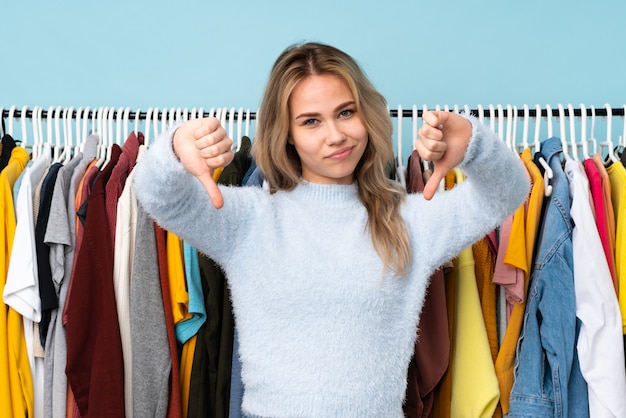 Menina russa adolescente comprando roupas na parede azul, mostrando o polegar para baixo