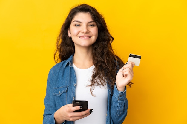 Menina russa adolescente isolada em fundo amarelo comprando com o celular com cartão de crédito