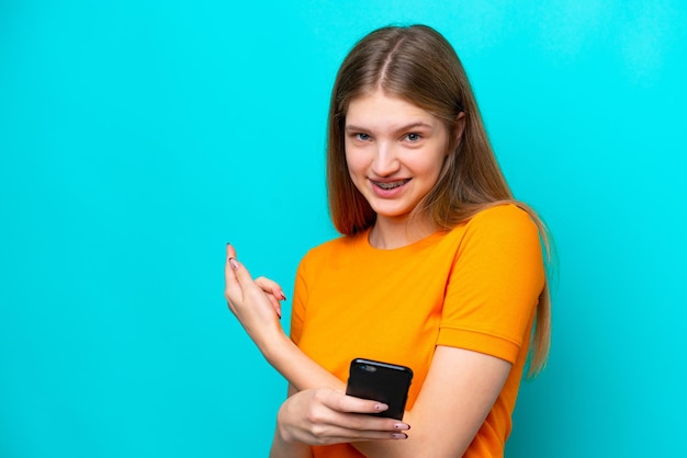 Menina russa adolescente isolada em fundo azul usando telefone celular e apontando para trás