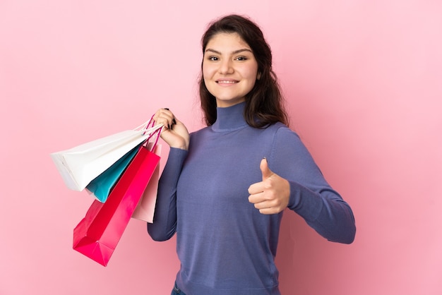 Menina russa adolescente isolada na parede rosa segurando sacolas de compras e com o polegar para cima