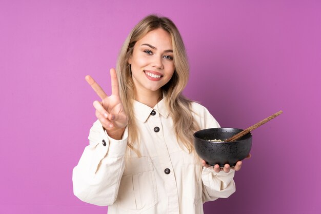 Menina russa adolescente isolada na parede roxa sorrindo e mostrando o sinal da vitória, segurando uma tigela de macarrão com pauzinhos