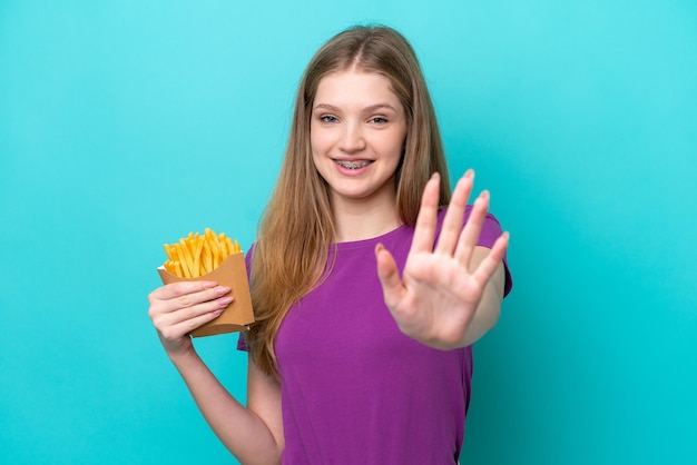 Menina russa adolescente pegando batatas fritas isoladas em fundo azul saudando com a mão com expressão feliz