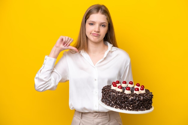 Menina russa adolescente segurando o bolo de aniversário isolado em fundo amarelo, mostrando o polegar para baixo com expressão negativa