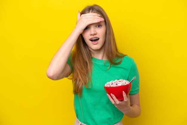 Menina russa adolescente segurando tigela de cereais isolada em fundo amarelo fazendo gesto surpresa enquanto olha para o lado