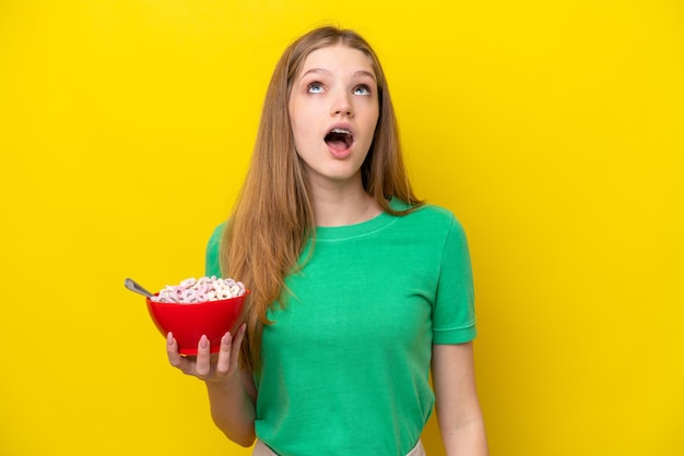 Menina russa adolescente segurando tigela de cereais isolada em fundo amarelo olhando para cima e com expressão de surpresa