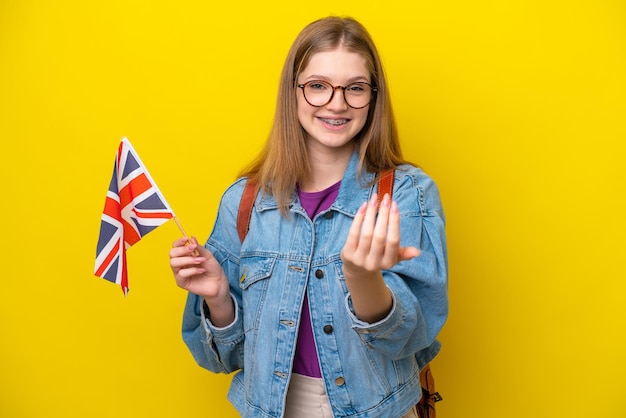 Menina russa adolescente segurando uma bandeira do Reino Unido isolada em fundo amarelo convidando para vir com a mão Feliz por você ter vindo