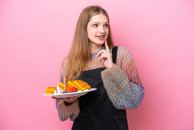 Menina russa adolescente segurando waffles isolados em fundo rosa pensando em uma ideia enquanto olha para cima