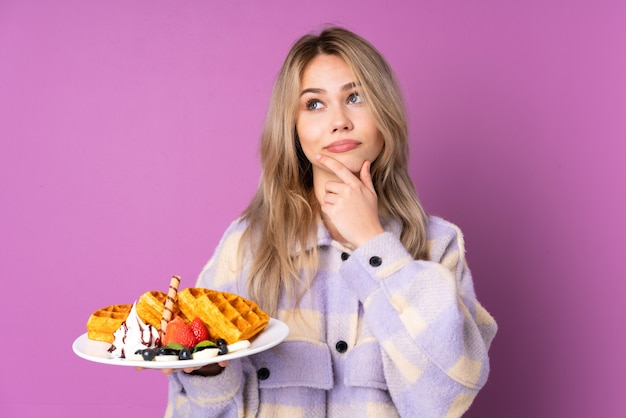 Menina russa adolescente segurando waffles isolados na parede roxa, tendo dúvidas e com expressão facial confusa