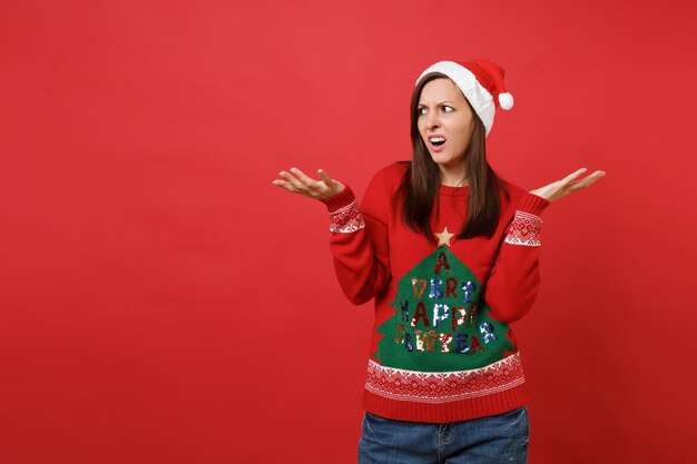 Menina Santa enojada irritada no suéter de malha, chapéu de Natal espalhando as mãos, olhando de lado isolado sobre fundo vermelho. Conceito de festa de feriado de celebração de ano novo feliz 2019. Simule o espaço da cópia.