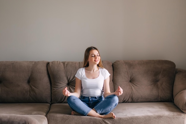 Menina saudável e serena meditando com os olhos fechados sentada no sofá em casa em posição de lótus jovem consciente relaxando fazendo ioga