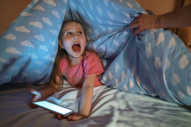 Menina, se escondendo sob o cobertor com smartphone à noite, quando todo mundo está dormindo