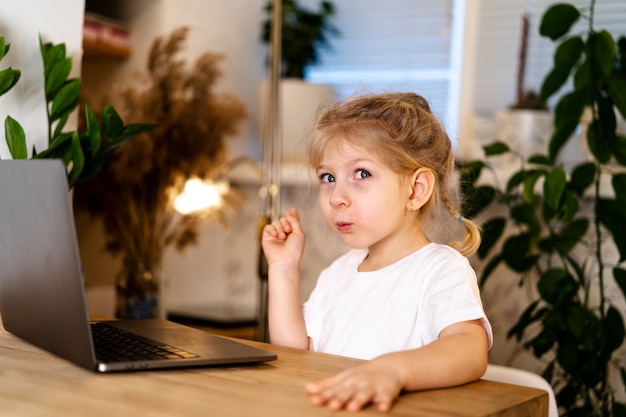 menina se senta a uma mesa com um laptop com o dedo para cima, sorri e fica esperta, experimentando