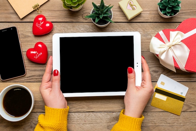 Menina segura tablet, cartão de débito, escolhe presentes, faz compras, xícara de café, dois corações, bolsa na mesa de madeira