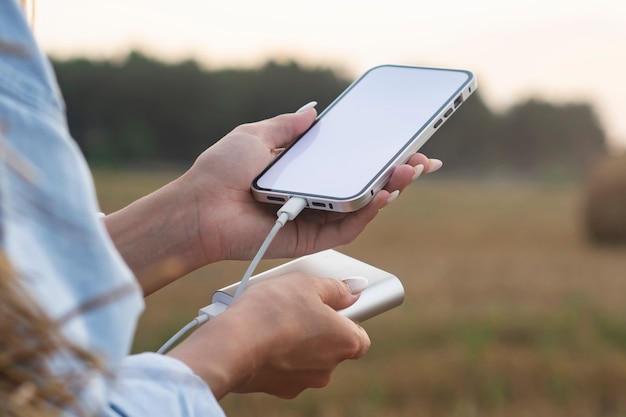 Menina segura uma maquete de um smartphone com uma tela branca nas mãos. Power Bank carrega o telefone contra o fundo da natureza.