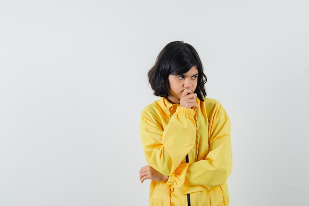 Menina segurando a mão no queixo com capuz amarelo e olhando pensativa, vista frontal.