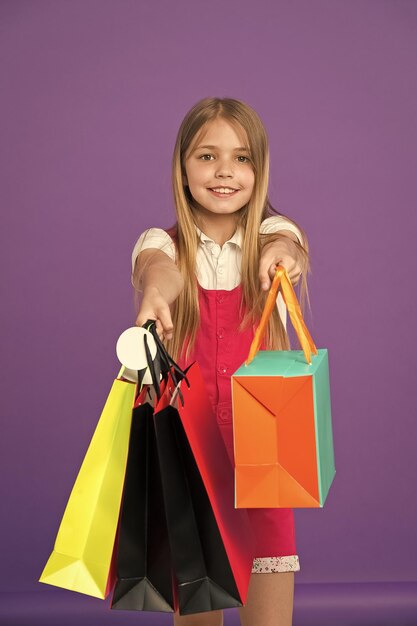 Foto menina segurando sacolas de compras coloridas modelo com longos cabelos loiros em fundo violeta criança dando presentes em sacos de papel clima de férias pequeno consumidor viciado em compras moda e conceito de vendas