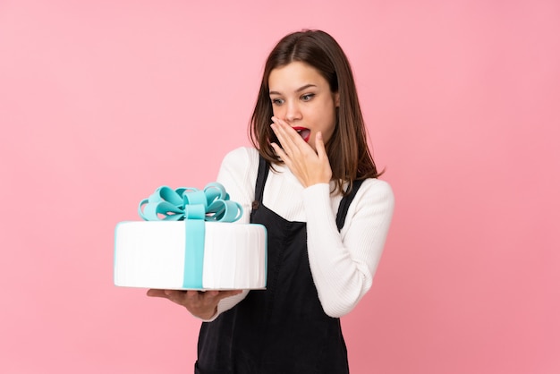 Menina segurando um bolo grande na parede rosa com expressão facial surpreso e chocado