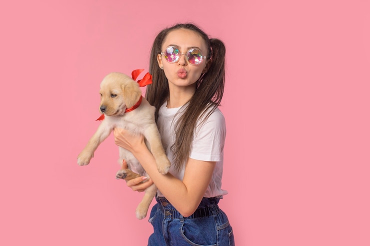 Foto menina segurando um cachorrinho com uma fita vermelha nos braços sorriso e felicidade em um fundo rosa